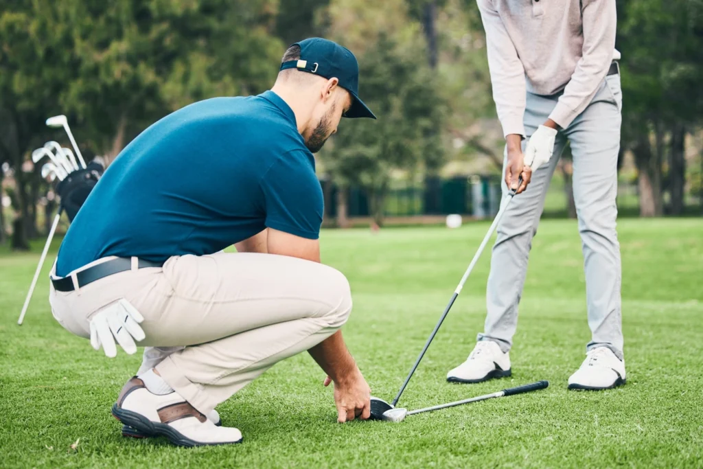 Golf instructor crouching on the grass to assist a student with their stance and club position during a lesson on a golf course.