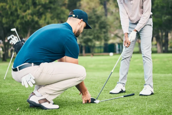 Golf instructor crouching on the grass to assist a student with their stance and club position during a lesson on a golf course.