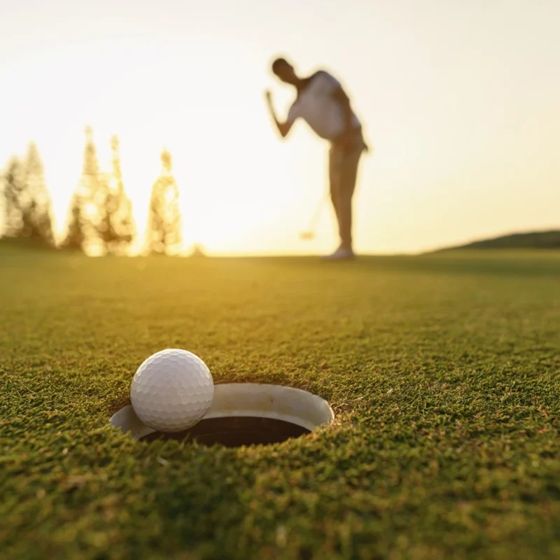 Close-up of a golf ball on the edge of the hole with a golfer celebrating in the background at sunset on a green course.
