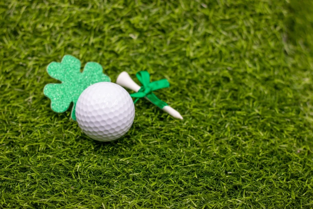 Golf ball on grass beside a green glitter shamrock and a white tee tied with a green ribbon, symbolising Irish-themed golf.