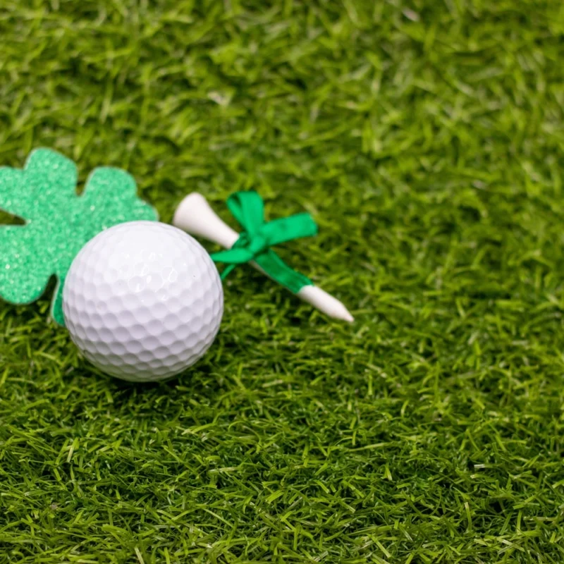 Golf ball on grass beside a green glitter shamrock and a white tee tied with a green ribbon, symbolising Irish-themed golf.