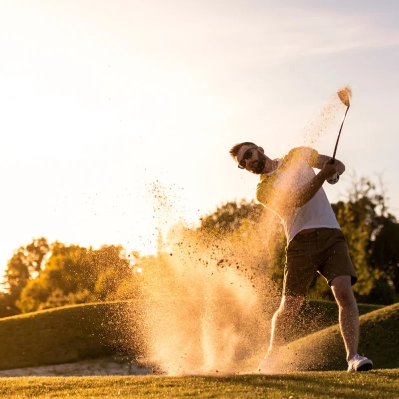 Man playing golf at sunset, swinging from a sand bunker as sand sprays into the air.