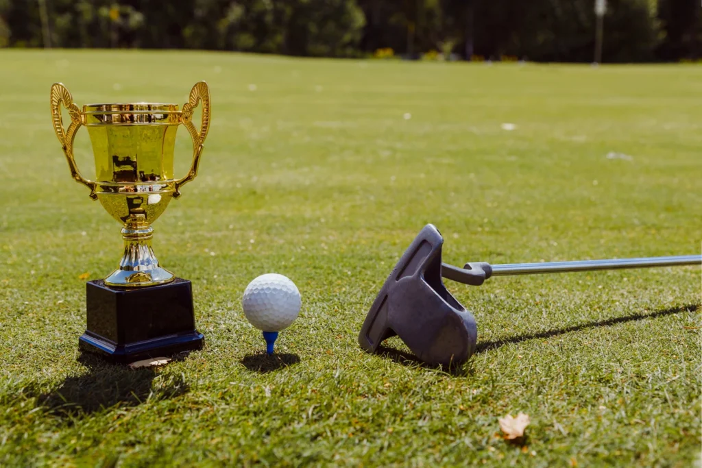 A close-up of a golf trophy, golf ball on a blue tee, and a putter resting on the green under bright sunlight.