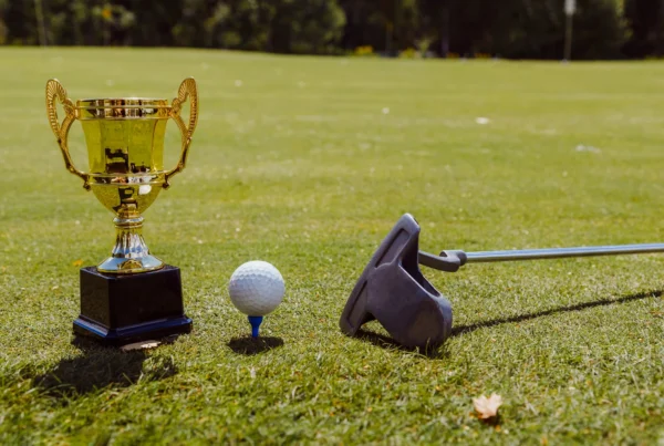 A close-up of a golf trophy, golf ball on a blue tee, and a putter resting on the green under bright sunlight.