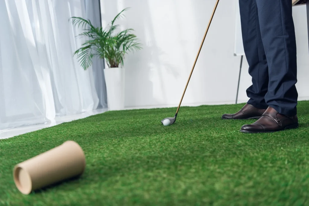 A person practising golf indoors on a green mat, preparing to hit a golf ball with a club, with a plant and window light in the background.