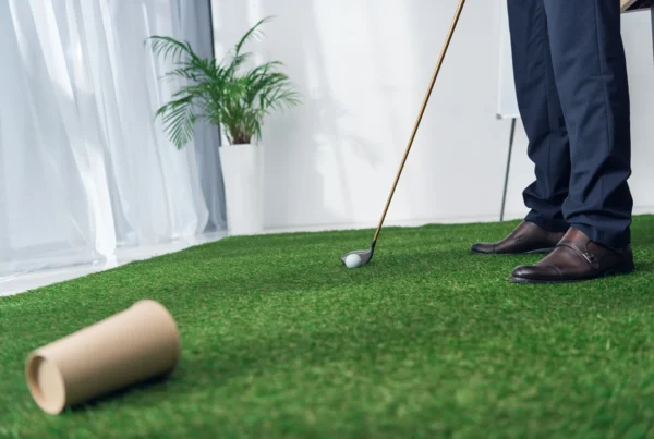 A person practising golf indoors on a green mat, preparing to hit a golf ball with a club, with a plant and window light in the background.
