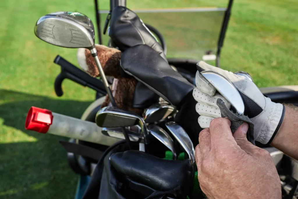 Golfer cleaning an iron club while standing beside a golf bag filled with clubs, headcovers, and accessories.