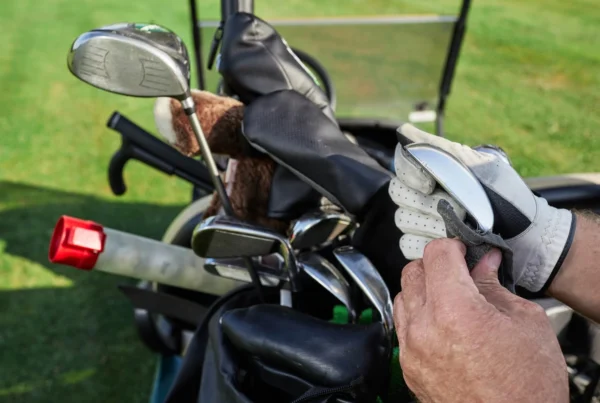 Golfer cleaning an iron club while standing beside a golf bag filled with clubs, headcovers, and accessories.