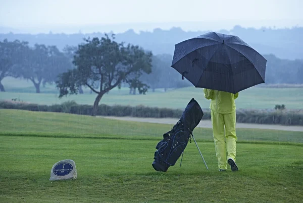 Golfer standing on a wet tee box in heavy rain with an umbrella and golf bag, reflecting typical winter conditions that lead to Winter Rules and WHS score restrictions.